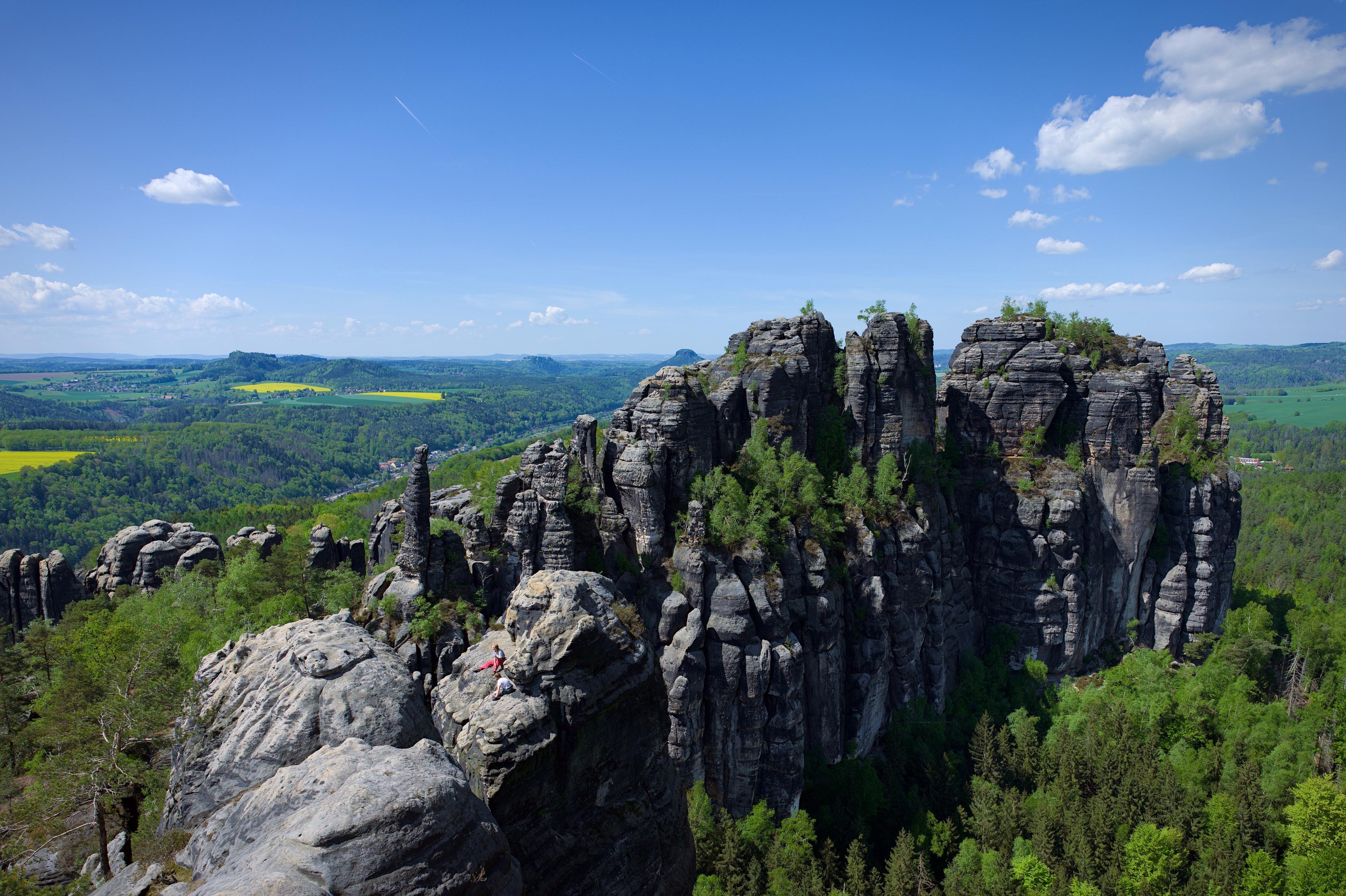 This photo shows the dramatic rock formations of the Elbe Sandstone Mountains in Saxon Switzerland National Park, Germany. Towering, weathered sandstone cliffs rise from a lush green forest, with scattered trees even growing from crevices in the rock. The sky is a rich blue with scattered cumulus clouds and faint contrails, suggesting clear spring or early summer weather.
In the foreground, two rock climbers rest on a large boulder, adding a sense of scale to the immense landscape. Behind them, the rugged vertical cliffs and pillar-like rock formations dominate the scene, resembling natural fortresses. The background stretches into rolling hills and patchwork fields, giving a sense of depth and openness. This photo shows the dramatic rock formations of the Elbe Sandstone Mountains in Saxon Switzerland National Park, Germany. Towering, weathered sandstone cliffs rise from a lush green forest, with scattered trees even growing from crevices in the rock. The sky is a rich blue with scattered cumulus clouds and faint contrails, suggesting clear spring or early summer weather.
In the foreground, two rock climbers rest on a large boulder, adding a sense of scale to the immense landscape. Behind them, the rugged vertical cliffs and pillar-like rock formations dominate the scene, resembling natural fortresses. The background stretches into rolling hills and patchwork fields, giving a sense of depth and openness.