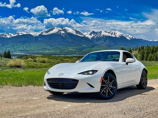 A white Mazda MX-5 Miata RF parked on a dirt turnout in front of a panoramic mountain range in Colorado. Snow-capped peaks rise in the background under a vivid blue sky with scattered clouds.