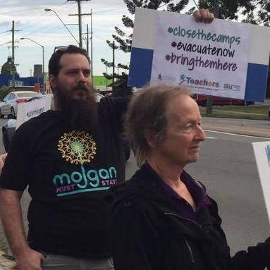 A group of protesters stand on a suburban roadside, holding signs aloft in a show of solidarity. The atmosphere is charged with purpose. In the foreground, you can see me wearing a black t-shirt emblazoned with a radiant, circular design and the words “mojgan must stay” standing firmly, one arm raised high to display a large placard. The sign is boldly lettered with hashtags #closethecamps, #evacuatenow, and #bringthemhere, calling for urgent action on refugee and asylum seeker issues. Another protester stands nearby, dressed in a dark jacket, gripping their own sign. Behind them, more demonstrators line the curb, their signs partially visible. The street is bordered by a row of trees and power lines, and the sky is overcast, lending a sombre tone to the scene. The group’s presence outside Peter Dutton’s office in the seat of Dickson underscores their demand for justice and compassion in Australia’s immigration policies.