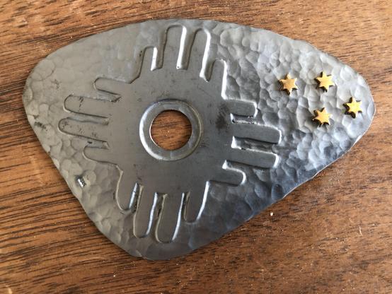 A hammered, blackened metal piece shaped like a shield with a central hole, featuring a bold application and four small gold stars on one side. The item rests on a wooden surface.