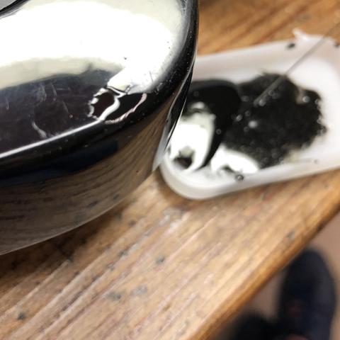 A close-up image of a shiny black object, possibly a guitar, with a wet reflection visible on its surface. Next to it is a small white container holding a dark, viscous substance and an applicator tool. The background shows a wooden surface.