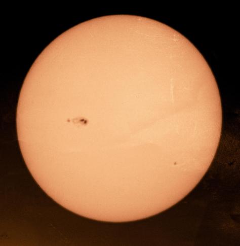 An orange colourised image of the face of the Sun as projected onto white card through binoculars, with a large sunspot group very easily visible and another smaller one on the opposite side. Photographed with my phone.
