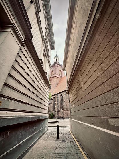 Ultraweitwinkelaufnahme. Eine schmale Kopfsteinpflastergasse zwischen zwei Gebäuden führt zu einer Backsteinkirche mit einem hohen Turm und einem spitzen Dach. Die Gebäude haben strukturierte Wände und der Himmel ist bewölkt.