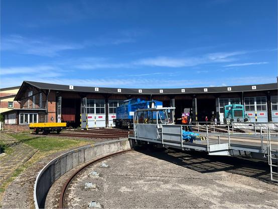 Drehscheibe, dahinter der Lokschuppen, vor dem ein kleiner gelber Güterwagen, eine blaue Diesellok der Pressnitztalbahn und eine türkisfarbene Rangierlok geparkt sind.