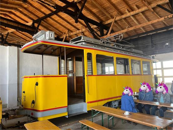 Gelber Straßenbahnwagen Tw 31 mit drei roten Streifen an der Seite (unten, mittig und oben) und offenen Bühnem an beiden Seiten, im Lokschuppen stehend. Davor Bierzeltgarniuren, an einem Tisch sitzen drei Personen. Das Dach sieht so aus, als sei es relativ neu gedeckt worden.
