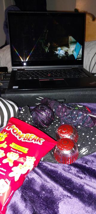 A bunch of stuff on a purple fleece blanket on my bed. In the foreground is a bag of Pom Bear crisps, two Tunnock's Tea Cakes, and some knitting on and next to a project bag made from a stars and planets print fabric. Behind that is a laptop showing an episode of What We Do In The Shadows. There is a rolled up grey weighted blanket behind that, and the edge of a black and white striped cushion visible on the left.