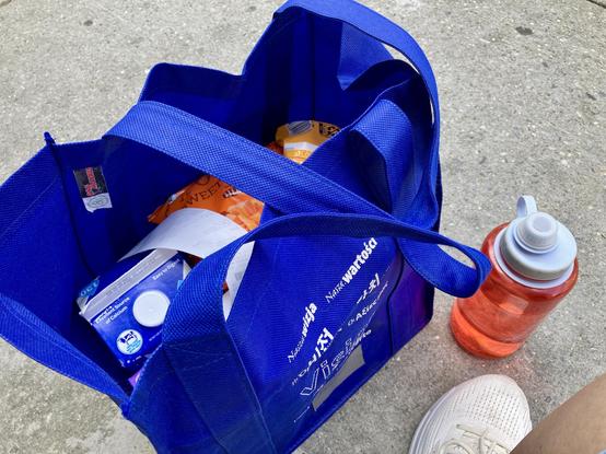 A blue reusable shopping bag filled with groceries, including a carton of milk and frozen veggies, rests on a concrete surface. Next to the bag is an orange water bottle filled halfway, and a white sneaker is partially visible.