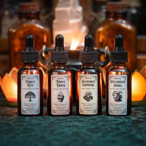 Four small dropper bottles of Beard Oil Set sit in a line. Their labels list their ingredients. The first bottle is labeled Forest Rite; the second is Temple Ember; the third reads Alchemist Grimoire; and the last is Spellbound Smoke. These are exclusive products from Inked Goddess Creations.