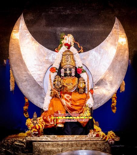 Idol of Hindu deity Krishna dressed in Orange, seated with left leg folded and over the right knee. A silver disc (emulating the Sun) and a large silver halfmoon behind Krishna. Krishna wears gold ornaments and a gold crown. White flower garland falls from the crown on to either side of Krishna. Blue cloth background.  