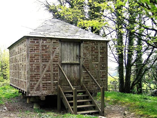 A small building set on mushroom-shaped stones, with walls of brick and wood and a slate roof. There is a wooden door on one side with a flight of wooden steps leading up to it.