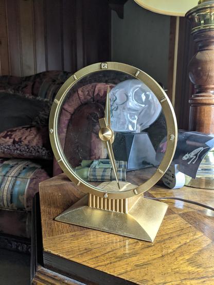Photo of a Jefferson Golden Hour clock I found at a rummage sale today. It's sitting on an end table plugged in to test it near the window. It's a round clock with a clear glass face, the outline, hands & base is a gold color.