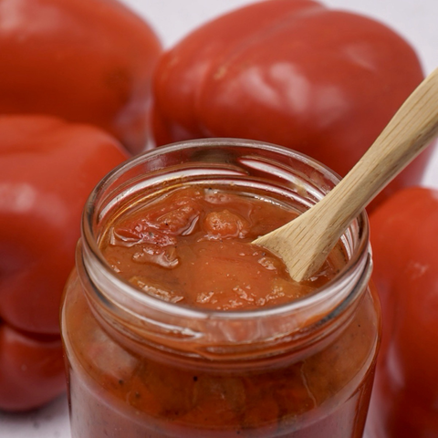 An open jar of red pepper chutney next to some red peppers. 