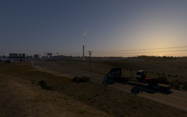 A scenic view from American Truck Simulator showing a rural area at dusk. The image features a road with a truck carrying a load, utility poles with power lines, and a distant industrial structure emitting smoke. The sky is transitioning from day to night, creating a serene atmosphere with the sun setting in the background. Sparse vegetation and a few buildings are visible along the roadside.