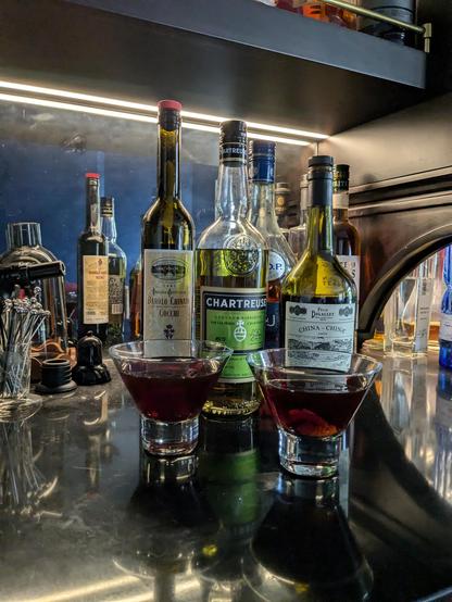 Two deep red cocktails in stemless cocktail glasses sit on a dark bar. Bottles of alcohol are behind the glasses: Barolo Chinato Cocci, Green Chartreuse, Bigallet China China amer and VSOP cognac. There are other things on the bar but they are not part of the drink.