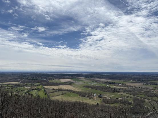 View of a vast plain from atop a mountain with Ottawa river in the background and a blue sky covered with white clouds for most of it 