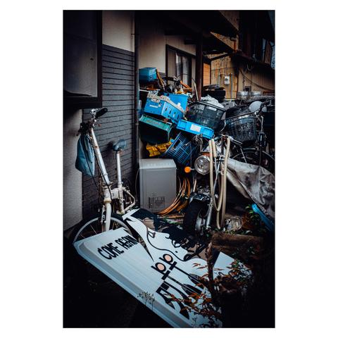 A narrow alley beside a residence is overtaken by a glorious avalanche of household castoffs—plastic crates, tangled wiring, broken bicycles, and a motorcycle peeking from beneath a tattered tarp.