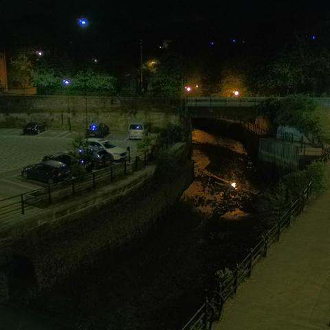 Photograph of the tidal stretch of Ouseburn River at south side of Ouseburn Bridge in Newcastle upon Tyne. To the left (south-west) of the bridge is Cut Bank, and to the right (north-east) is Byker Bank. In the foreground is the pedestrian Riverside Walk and the photo also spans some surrounding built-up areas and trees, with sky in the background. Still image captured from webcam at 03:13 hrs on Sunday 4 May 2025.