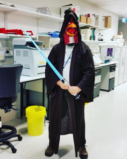 In a brightly lit biosecurity level 2 (BSL2) microbiology laboratory, I stand out against the backdrop of white countertops, shelves lined with lab equipment, and pops of colour from red trays and a vivid yellow clinical waste bin. 


I am dressed in a dramatic, floor-length Jedi robe of deep brown, its wide sleeves draping over my arms. Facing the camera and completing the ensemble, I wear a comically oversized rooster mask, its orange beak and red comb boldly jutting out, feathers painted in lively hues. 


In my hands, I grip a glowing blue lightsaber, holding it confidently at an angle in front of my body as if defending from errant StormTrooper fire.  


The playful fusion of Star Wars Day and International Respect For Chickens Day is unmistakable-your costume humorously bridges science fiction and animal advocacy, bringing a sense of fun and celebration into the serious, orderly environment of the laboratory. 


The scene is both whimsical and memorable, with my creative outfit injecting joy and personality into the sterile lab setting.