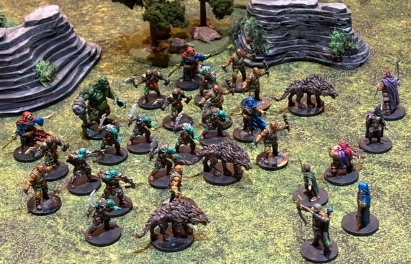 A photo of a wargame tabletop with forest and stone terrain. On the left are painted figurines of an ogre, orcs, skeletons, goblins, bandits, and wargs. In the right are six heroes - a hobbit, two elves, a dwarf, and two humans.