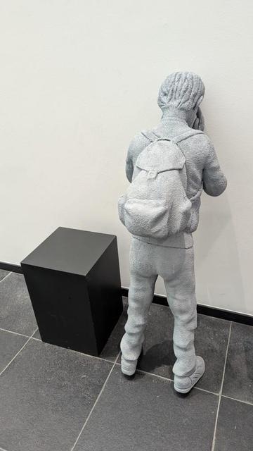 The photo shows a boy called Issam, a sculpture in the Skam Museum in Ghent. It is a refugee child, leaning its head against the wall and covering its eyes like in the children's game where you count and then look for your friends who are hiding. To the left is a black block the size of a stool.