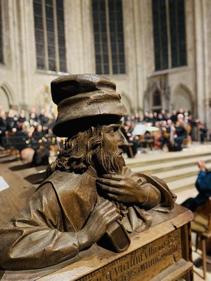 Gotischer Kirchenraum. Im Vordergrund eine geschnitzte Figur mit einem Buch in der einen Hand, streicht sich mit der anderen Hand den Bart. Voluminöser, gequetschter Hut. Dahinter unscharf Chorraum. In der erhöhten Apsis Chor und Orchester