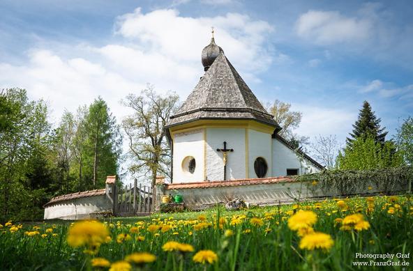 A serene outdoor scene featuring a small white building with a cross on top, set against a backdrop of lush green grass and a clear blue sky with fluffy white clouds. Surrounding the building are various plants and trees, adding to the peaceful atmosphere of the landscape. The building is the focal point, standing out with its bright white color and the prominent cross on its roof. This picturesque setting evokes a sense of tranquility and spirituality, making it a perfect representation of a peaceful countryside church or chapel nestled in nature.