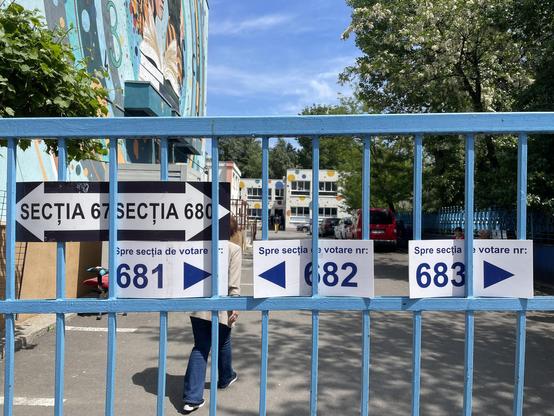 Gate with directional signs indicating polling sections 67, 680, 681, 682, and 683. A person is walking past, with a colorful mural visible on the wall behind and trees in the background.