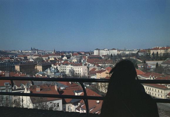 The image depicts a panoramic view of a city with a clear blue sky. In the foreground, a person is silhouetted against the cityscape, standing behind a railing, looking out over the rooftops. The city features a mix of architectural styles, with buildings having red-tiled roofs and some with white facades. The buildings are densely packed, with varying heights and designs, suggesting a historic urban area. In the background, there are prominent structures, including a large white building with a classical facade, and a castle-like structure with spires, indicating a significant historical site. The overall scene is bright and clear, with no visible clouds, suggesting a sunny day.
