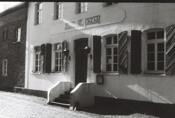A white stucco beer hall with wooden shutters along a cobblestone street. The sign on the front says "Zum St. Peter"