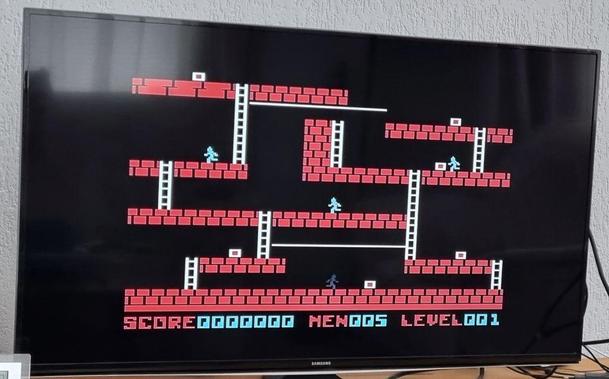 The first level of the game Lode Runner on a Samsung tv. The level is made up of different red platforms with white ladders connecting the platforms. A dark person-shaped sprite [player character] is seen on the bottom, three white [enemy] characters are on higher platforms. Spread out over the level are several white-and-read squares. Text at the bottom shows the current level info: "Score0000000 Men005 Level001