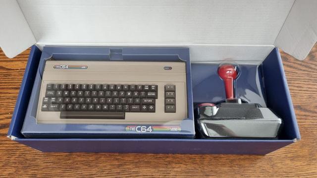 An oblong box that shows a mini replica of the Commodore 64 breadbox computer, next to a full size joystick similar to the Competition Pro.

Colors: the box is blue with a white cover, the C64 is beige with black an grey keys like the original, the joystick is black with a red grip. The box is seen on wooden background of different shades of brown.
