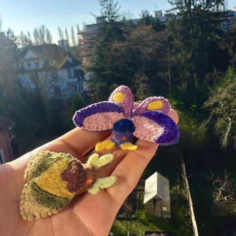Two little felt moths resting on my hand in front of a sunlit garden backdrop. 
One moth has its wings down and is pale yellow, green and brown. The other has open pink and purple wings and body, yellow antennas and yellow spots on its wings. 