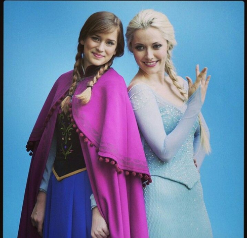 A imagem mostra Anna e Elsa, interpretadas por Elizabeth Lail e Georgina Haig, respectivamente, na série Once Upon a Time. Elas estão juntas, posicionadas lado a lado, contra um fundo azul claro. Anna está à esquerda, usando um vestido azul escuro com um casaco roxo-magenta com detalhes em pompons. Seu cabelo castanho-avermelhado está trançado em duas tranças. Ela está sorrindo gentilmente, com uma expressão calorosa e amigável. Elsa está à direita, usando um vestido longo e elegante de cor azul-claro gelo, com mangas compridas. Seu cabelo loiro está trançado em uma única trança que cai sobre um ombro. Ela também está sorrindo, mas com uma expressão um pouco mais reservada que a de Anna. Ambas as personagens parecem estar em um estúdio fotográfico, com o fundo liso e sem detalhes. A imagem como um todo transmite uma sensação de irmandade e união entre as duas personagens.