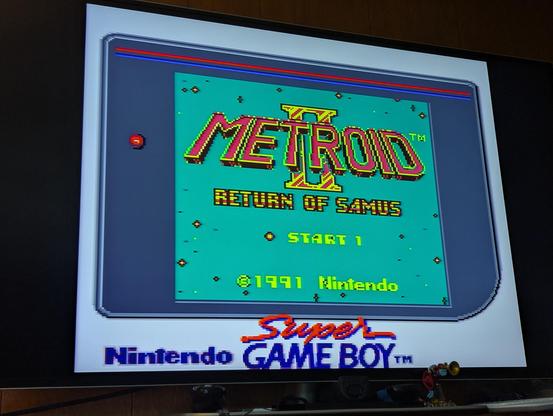 A photograph of a flat-panel television, displaying the title screen of "Metroid II: Return of Samus" being run on a Super Nintendo using a Super Game Boy adapter.