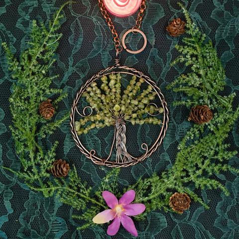 An example of the Peridot Tree of Life Altar Decor Hanging is laid flat on a lace-covered surface. Twisted and braided antiqued copper wire make the circular frame and tree trunk. Small peridot chip beads represent the leaves. The frame hangs from a copper chain ending in a copper split ring. Presented by Inked Goddess Creations.