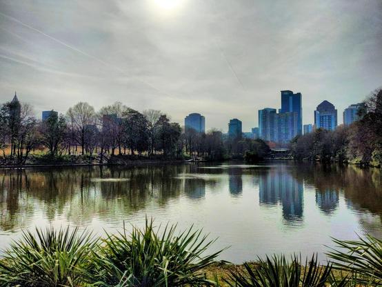 A calm lake in a city park with trees along the shore and tall buildings in the background. The water reflects the skyline and trees. The sky is hazy with the sun faintly shining.