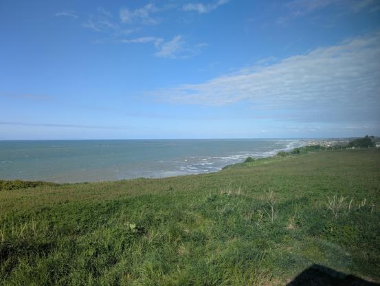 Wir blicken über eine grasbewachsene Fläche auf das Meer, das in verschiedenen Blautönen zu sehen ist. Die Gischt der Wellen ist weithin zu sehen. Im Osten liegt ein Ort mit vorgelagertem Sandstrand am Wasser.