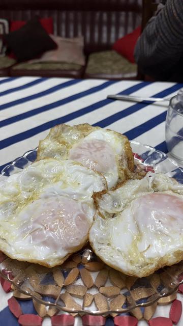 Three fried goose eggs with soft yolks, on a glass plate, placed on a table with a blue and white striped tablecloth. Behind, we can see a sofa with pillows in different colors. It looks like a cozy and homey place.