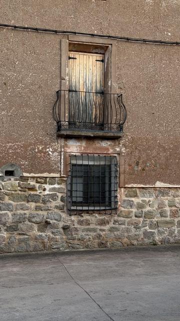 Wall of a rustic house with stone base and brown plaster. There is a window with a black metal grid and a small wooden door above with a curved iron balcony. Everything looks old and traditional, like in a small village.