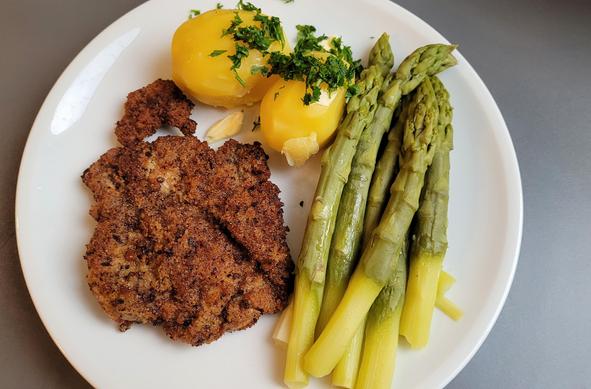 Farbfoto: Auf einem weißen Teller liegen ein paniertes Schnitzel, zwei Kartoffeln mit Petersilie und eine Portion grüner Spargel. 