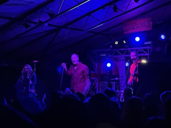 A blue and purple tinged photo of Jon King singing, flanked by Gail Greenwood on bass and Ted Leo on guitar. 
