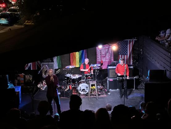 The whole band, with drummer Hugo Burnham now visible. Behind them, various flags including UK,US, pride, and a woman’s right to choose. 