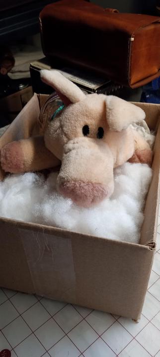 A small box with a plush toy pig inside it surrounded by white stuffing material.