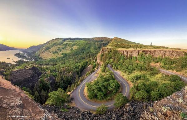 A winding road curves through a lush, hilly landscape with a river visible in the background under a clear sky.