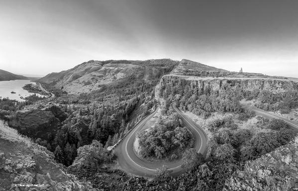 A winding road snakes through a lush mountainous landscape under a clear sky, with a river visible on the left.