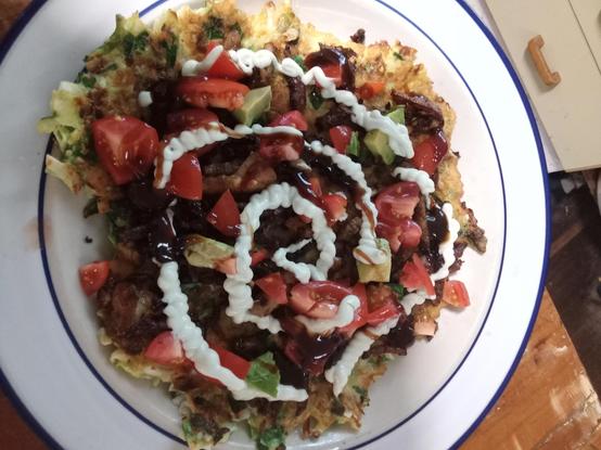A white plate with a blue tim, with a large cabbage pancake, red tomatoes avocado, and a spiral of mayo on top.