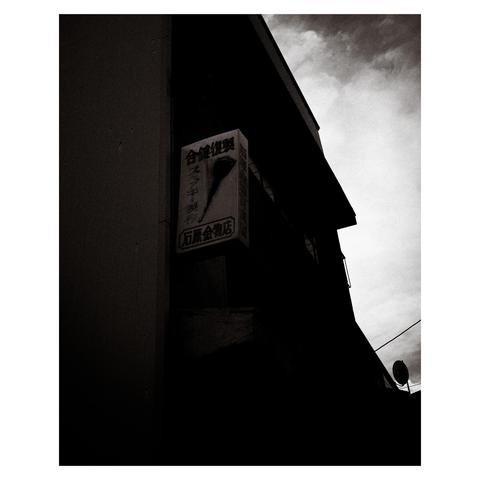 A vertical sign juts out from the dark facade of a narrow building, lit by faint skyglow. The sign features a stylized key and bold kanji reading 合鍵作製 (“duplicate key services”), with the business name 石原金物店 below. The surroundings fade into shadow, with only slivers of rooftop and power lines visible. 
