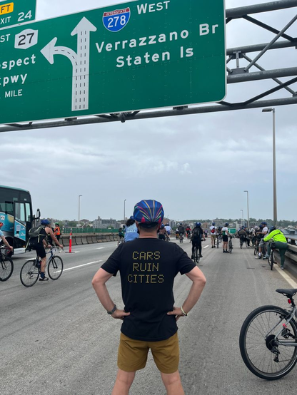 Person standing on highway with lots of bikes around him. He wears a bike helmet and a black t-shirt that reads "Cars Ruin Cities" on the back. He stands below a highway sign that says "278 Verrazzano Br Staten Is."
