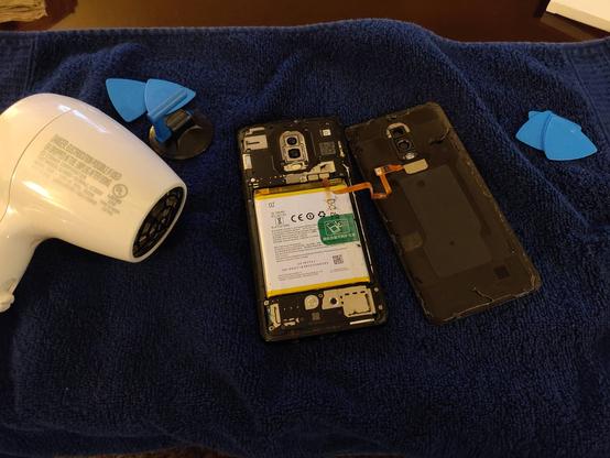 OnePlus 6 with the back cover removed, a hair dryer is sitting to the left of the phone.  iFixit opener pics and suction cup are around the phone.  There is a ribbon cable attached to the inside of the back cover for the fingerprint sensor that is still connected to the motherboard.
