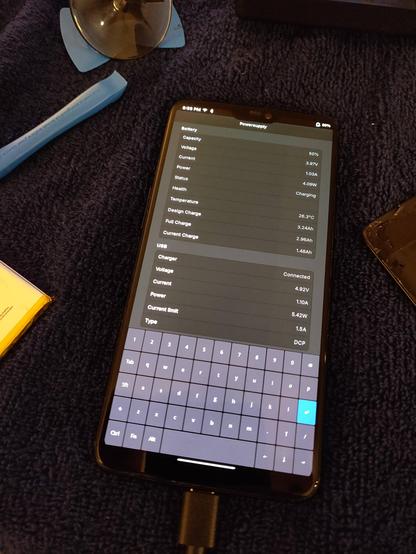 The phone, now powered up and booted into postmarketOS Phosh, with the Powersupply app open showing the battery charging at around 5W and the reported full capacity showing 2.96Ah.